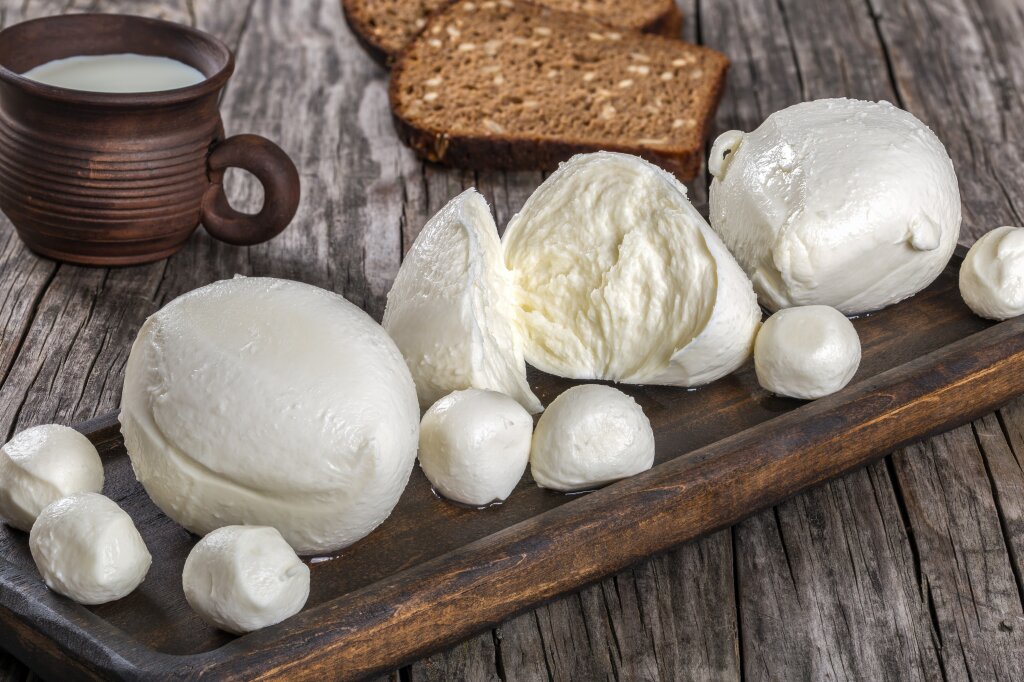 Fresh mozzarella balls and small mozzarella pearls on a wooden tray, with milk and bread alongside.